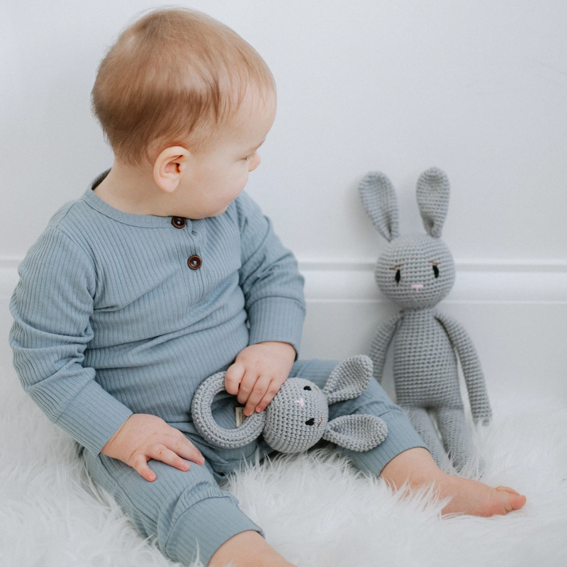 A baby wearing a blue long sleeve ribbed cotton romper sitting next to a gray knit rabbit toy and holding a knit rabbit rattle.
