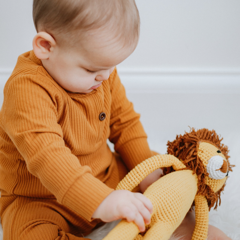 A baby wearing a long sleeve ribbed cotton romper in mustard colour, sitting and playing with a lion knit toy.