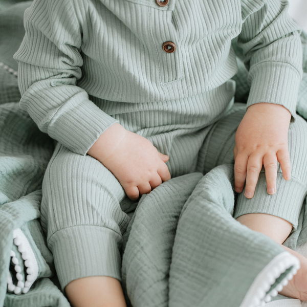 A baby wearing a long sleeve ribbed cotton romper in sage green color, with buttons on the front and popper fastenings at the leg.