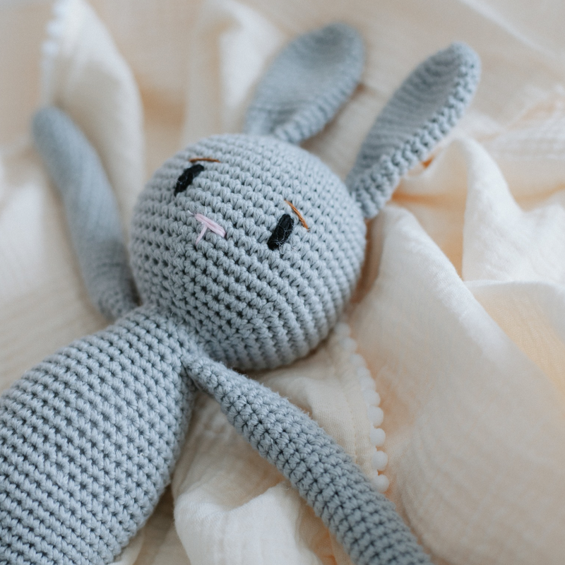 A handmade crochet grey bunny toy with a soft texture, laid on a white fabric surface.